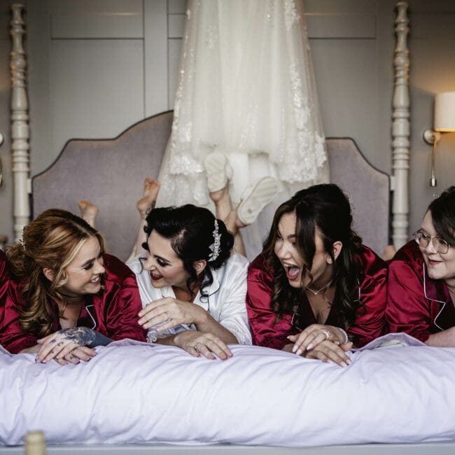 bridesmaids in red robes lying on the bed with wedding dress behind them before brides wedding day at sandburn hall hotel