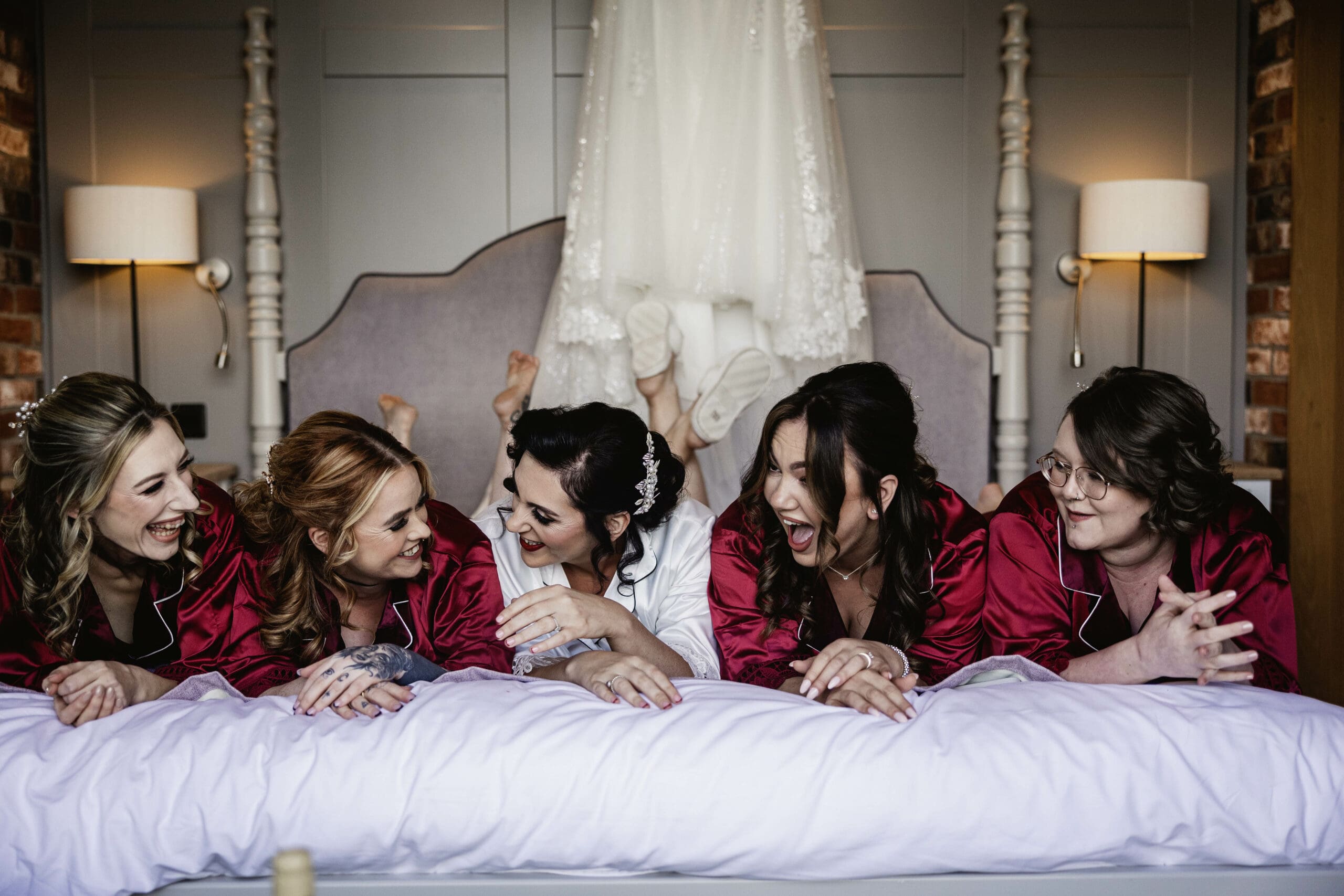 bridesmaids in red robes lying on the bed with wedding dress behind them before brides wedding day at sandburn hall hotel