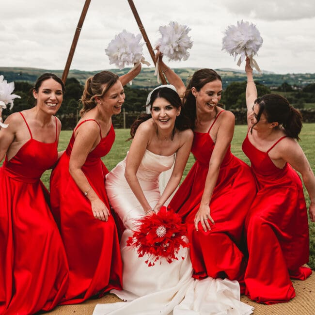 Bridesmaids in red outfits ducking down at east riddlesden hall holding white feathers in the air || West Yorkshire Wedding Photographer