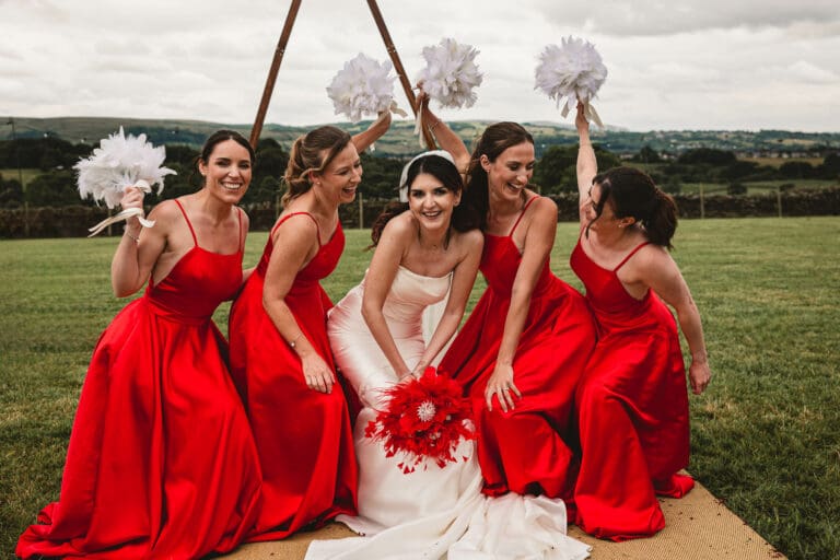 Bridesmaids in red outfits ducking down at east riddlesden hall holding white feathers in the air || West Yorkshire Wedding Photographer