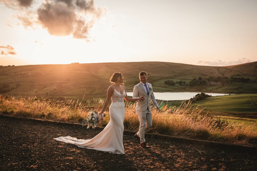 a golden sunset photo taken at saddleworh hotel as a bride and groom are walking with castleshaw lower reservoir in the background | Yorkshire wedding Photographer