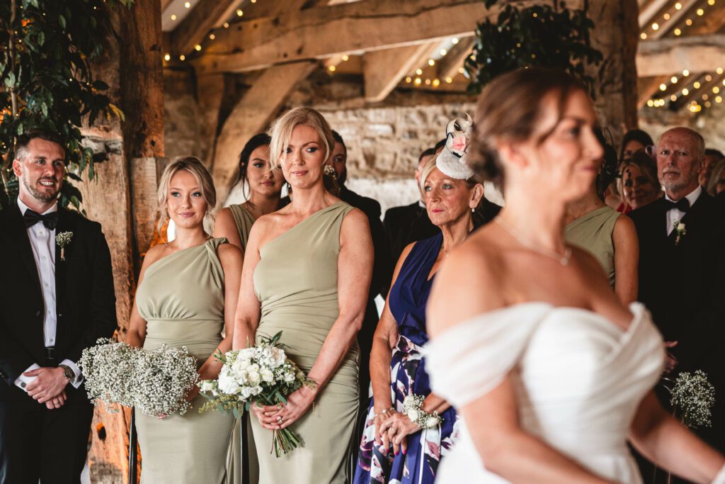 2 Bridesmaids and a bridesman look onwards to watch there friend getting married during the ceremony holding there flowers dressed in green sage bridesmaid addresses at the tithe barn