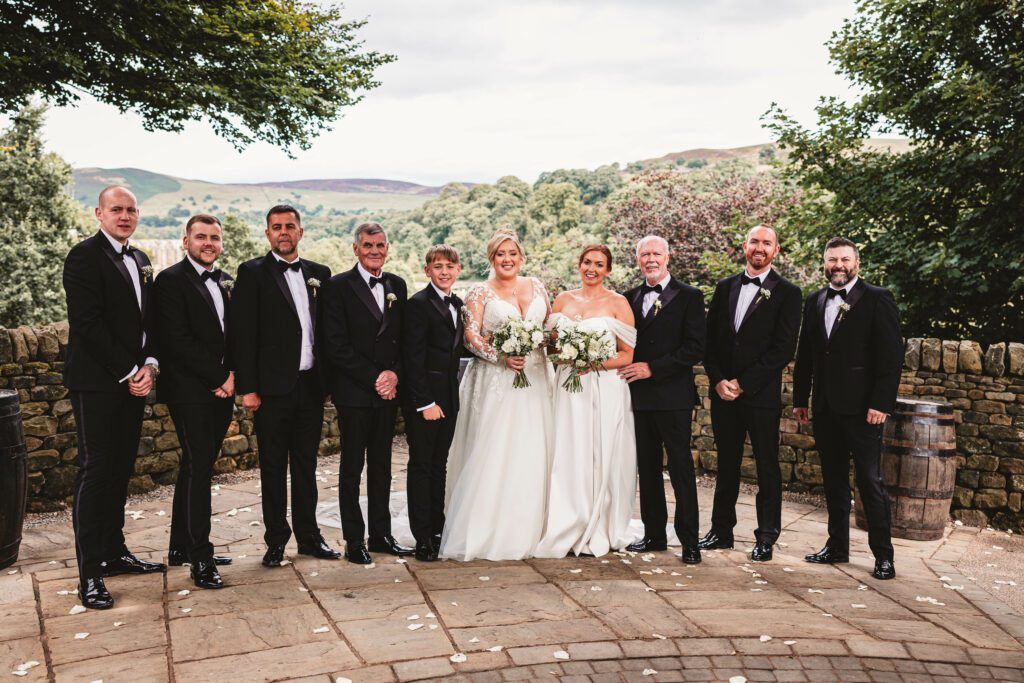 Groomsmen lined up taking a picture dressed in all black tuxedo outfits alongside the 2 beautiful brides with bolton abbey in the backaground at there wedding at the tithe barn