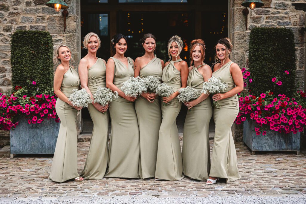 Bridesmaids dressed in sage green dresses holding white flowers stood infront of the tithe barn entrance