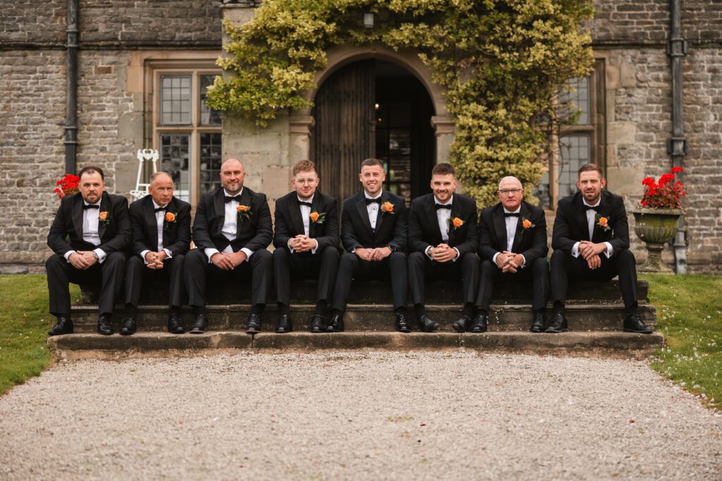 Groomsmen all lined up sat down dressed in black tuxedos with orange flowers at tissington hall | Tissington Wedding Phtographer